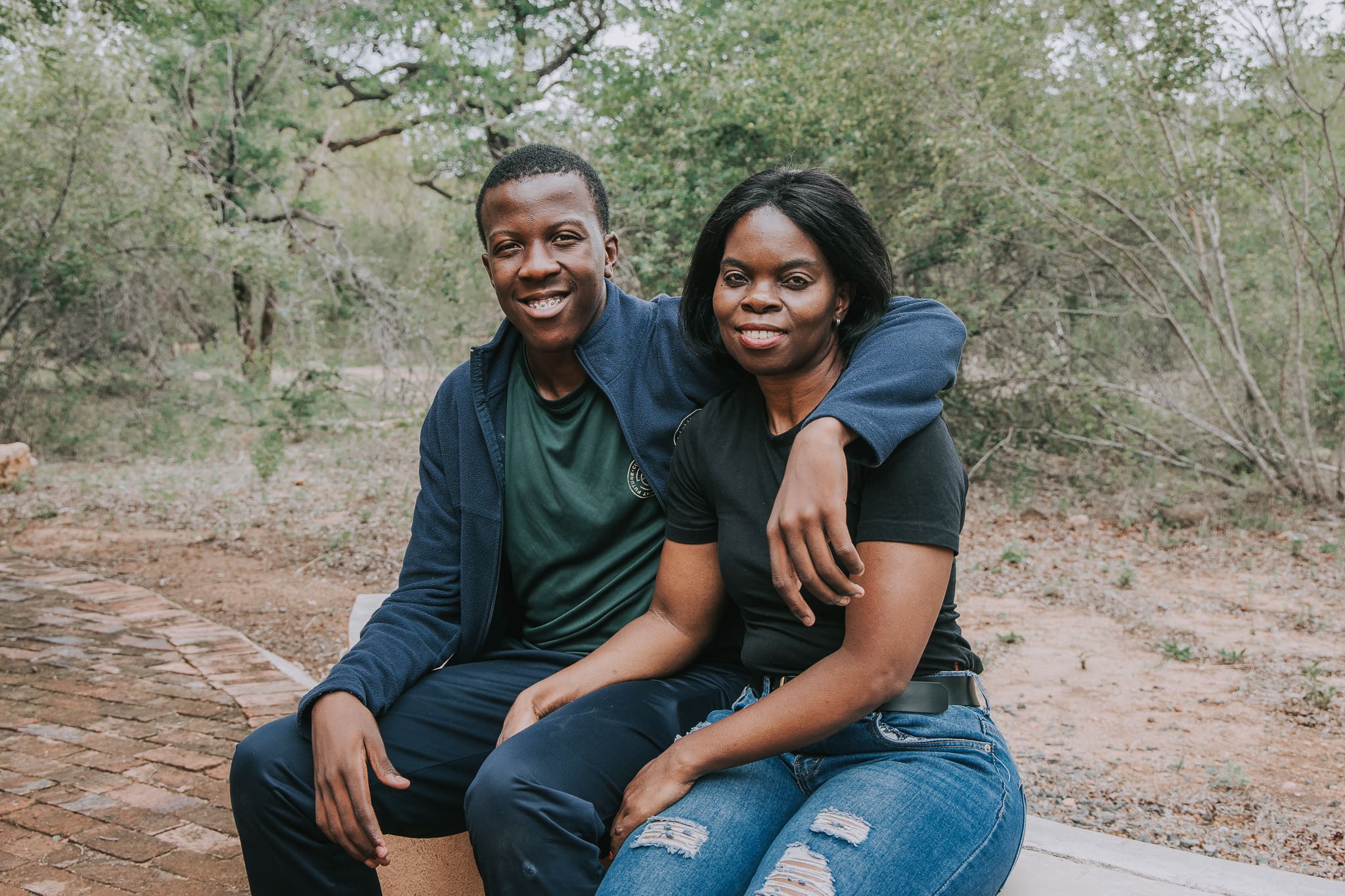 A young man and a woman sitting outdoors with his arm around her shoulder, both smiling.