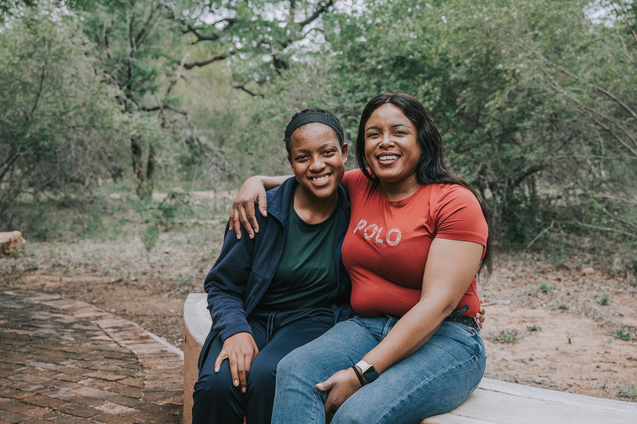 Two women sitting outdoors with arms around each other, smiling warmly at the camera.
