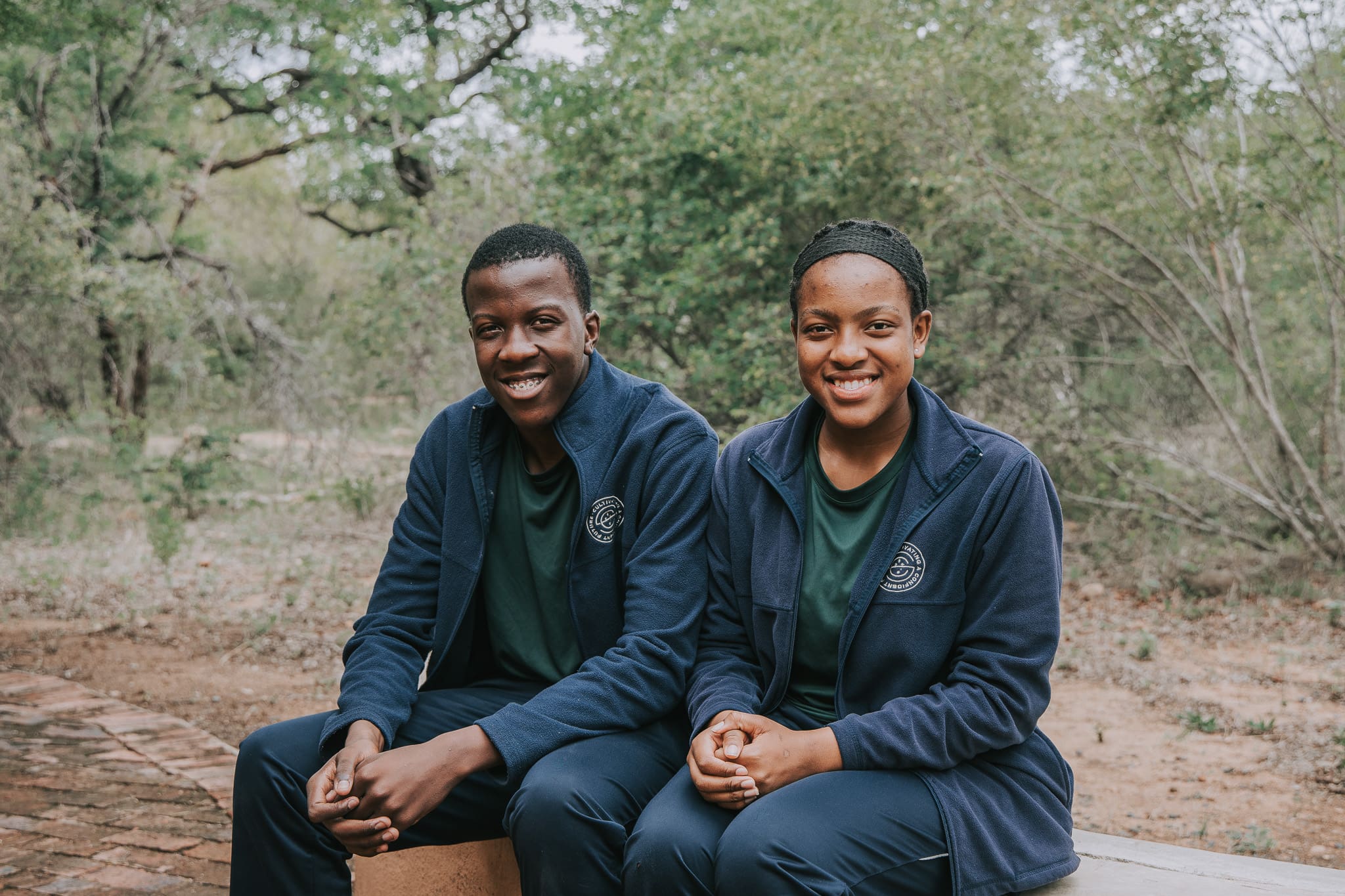 Two young Tanda Tula team members sitting outdoors in uniform, smiling toward the camera.